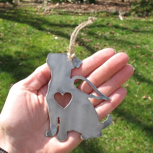May include: Metal ornament of a dog with angel wings and a heart cutout. The ornament is silver in color and has a rustic finish. It is hanging from a twine string.