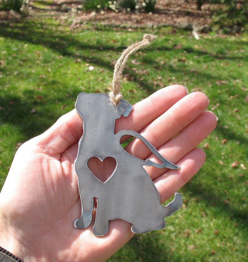 May include: Metal ornament of a dog with angel wings and a heart cutout. The ornament is silver in color and has a rustic finish. It is hanging from a twine string.
