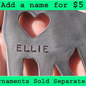 May include: A silver metal ornament shaped like a dog with a heart cut out. The ornament has the name "ELLIE" engraved on it. The text "Add a name for $5" and "Ornaments Sold Separately" are also visible in the image.