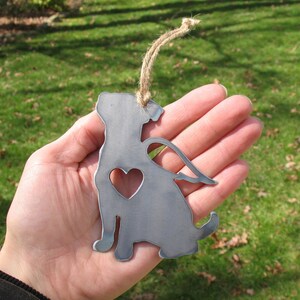 May include: Metal ornament of a dog with wings and a heart cutout. The ornament is silver and has a twine hanger.