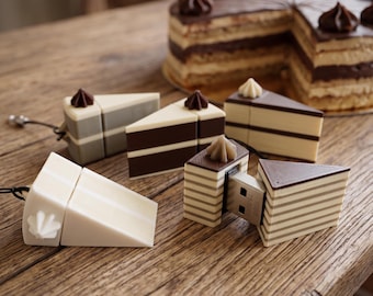 May include: A collection of USB flash drives shaped like triangular cake slices. The drives feature layered designs in shades of brown, cream, and white, topped with small frosting dollops. One drive is shown open, revealing the metal USB connector.