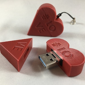Könnte beinhalten: Ein roter herzförmiger USB-Stick mit einem silbernen USB-Stecker. Der Stick ist geöffnet und der Stecker ist sichtbar. Der Stick ist außen mit "#LOVE" beschriftet. Der Stick liegt auf einer weißen Oberfläche.