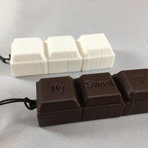 Könnte beinhalten: Zwei Schlüsselanhänger in Form von Schokoladentafeln, einer weiß und einer braun. Der weiße Schlüsselanhänger trägt die Worte "My Sweet". Der braune Schlüsselanhänger trägt die Worte "My Sweet" und ein Herz.
