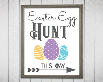 May include: A white sign with a gray frame and the text "Easter Egg Hunt This Way" in black. There is a gray arrow pointing to the right and three stylized eggs in purple, yellow, and blue.