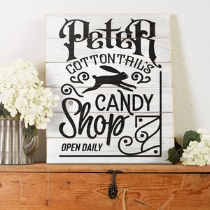 May include: White wooden sign with black lettering that reads "Peter Cottontail's Candy Shop" with a black silhouette of a rabbit running. The sign also says "Open Daily".