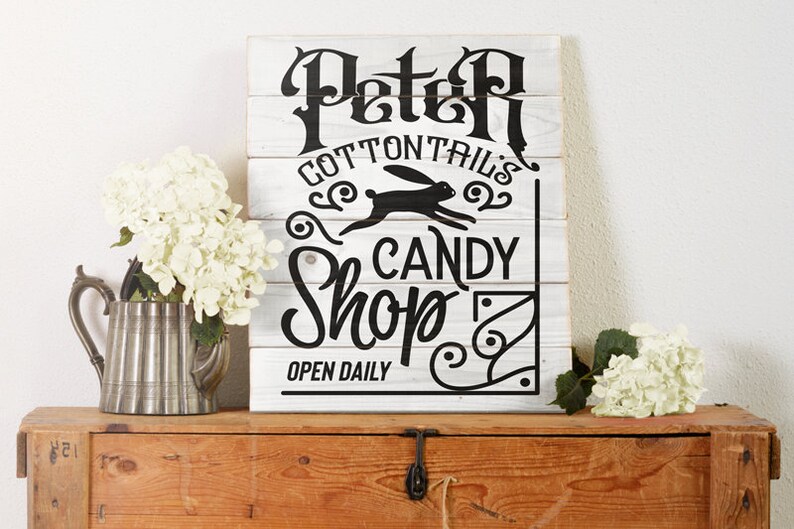 May include: White wooden sign with black lettering that reads "Peter Cottontail's Candy Shop" with a black silhouette of a rabbit running. The sign also says "Open Daily".