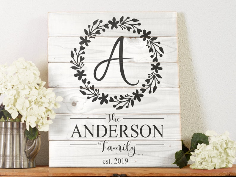 Op de afbeelding: Een wit houten bord met een zwarte bloemenkrans en de letter "A" in het midden. Onder de krans staat de tekst "The Anderson Family est. 2019".
