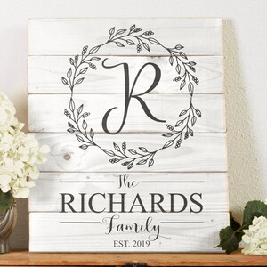 Peut inclure: Une pancarte en bois blanc avec une couronne florale noire et la lettre "R" au centre. Le texte "The Richards Family Est. 2019" est sous la couronne.