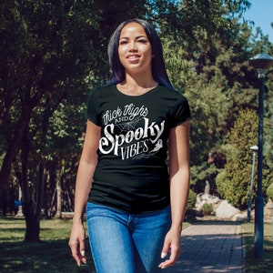 Puede incluir: Una camiseta negra con texto blanco que dice "Thick Thighs and Spooky Vibes". El texto est&aacute; en una fuente estilizada e incluye una telara&ntilde;a y un fantasma.