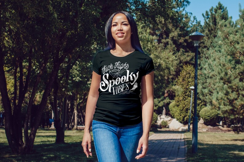 Puede incluir: Una camiseta negra con texto blanco que dice "Thick Thighs and Spooky Vibes". El texto est&aacute; en una fuente estilizada e incluye una telara&ntilde;a y un fantasma.