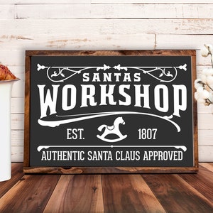 May include: Black and white sign with the text "Santa's Workshop" in a vintage style font. The sign also includes the text "Est. 1807" and "Authentic Santa Claus Approved".
