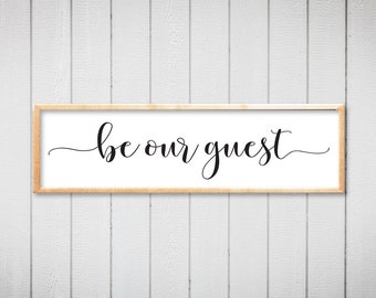 May include: A framed sign with the text "be our guest" in a cursive font. The sign is on a white wood background.
