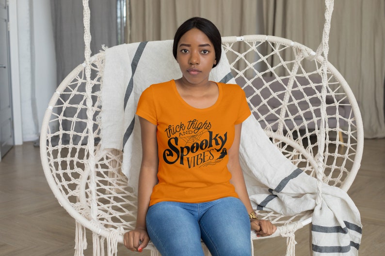 Puede incluir: Una mujer lleva una camiseta naranja con texto negro que dice "Thick Thighs and Spooky Vibes". El texto est&aacute; en una fuente estilizada con una telara&ntilde;a y un fantasma.