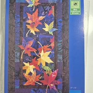 May include: A quilt pattern featuring colorful maple leaves in shades of red, orange, yellow, and green. The design includes a blue background and a dark brown border. The dimensions are 27 x 52 inches.
