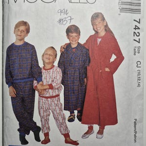 May include: McCall's 7427 sewing pattern envelope featuring illustrations of children wearing pajamas and robes. The garments include a blue plaid long-sleeve set, a white and red patterned set, a blue plaid robe, and a solid red long robe.