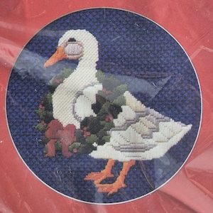 May include: Cross-stitch artwork of a white goose wearing a wreath with a red bow. The goose has an orange beak and feet. The background is a dark blue, and the artwork is framed in a red border.
