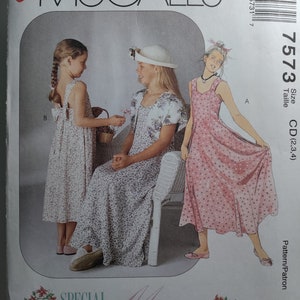 May include: A sewing pattern for two girls' dresses. The pattern is called "Easy McCall's" and the pattern number is 7573. The dresses are both sleeveless and have a floral print. The dress on the left is a longer, flowing style, while the dress on the right is shorter and more fitted. The pattern is for sizes CD (2, 3, 4).