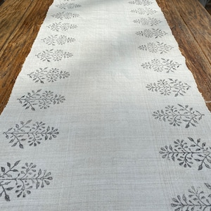 Könnte beinhalten: Ein weißer Tischläufer aus Leinen mit einem schwarzen Blumenmuster. Das Muster wiederholt sich über die gesamte Länge des Läufers.