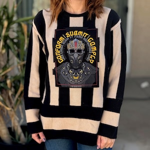 May include: A long-sleeve knit sweater featuring vertical black and beige stripes. The center displays a large rectangular graphic patch with a stylized robotic skull figure and the text "CONFORM SUBMIT COMPLY" in bold yellow lettering.