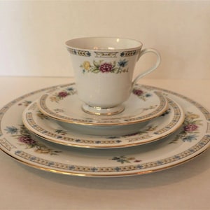 May include: A set of four white porcelain plates with a gold trim and floral pattern. The plates are stacked on top of each other, with a matching teacup and saucer on top.