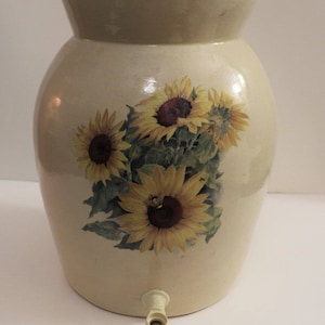 May include: A ceramic water dispenser with a spigot, decorated with a floral design featuring sunflowers in shades of yellow, brown, and green.