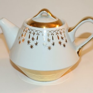 May include: White teapot with a gold rim and a gold design of stars and hanging ornaments. The teapot has a lid and a handle.