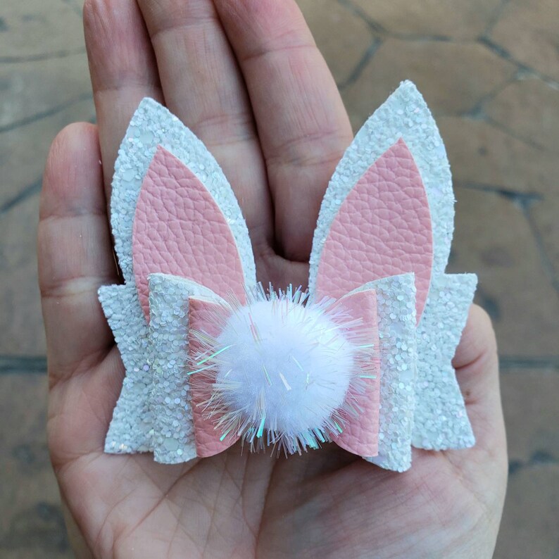 May include: A white glitter bunny ear hair bow with a pink faux leather bow and a white pom-pom in the center.