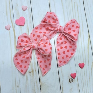 May include: Two pink hair bows with a heart pattern. The bows are tied together and are on a white wooden background.