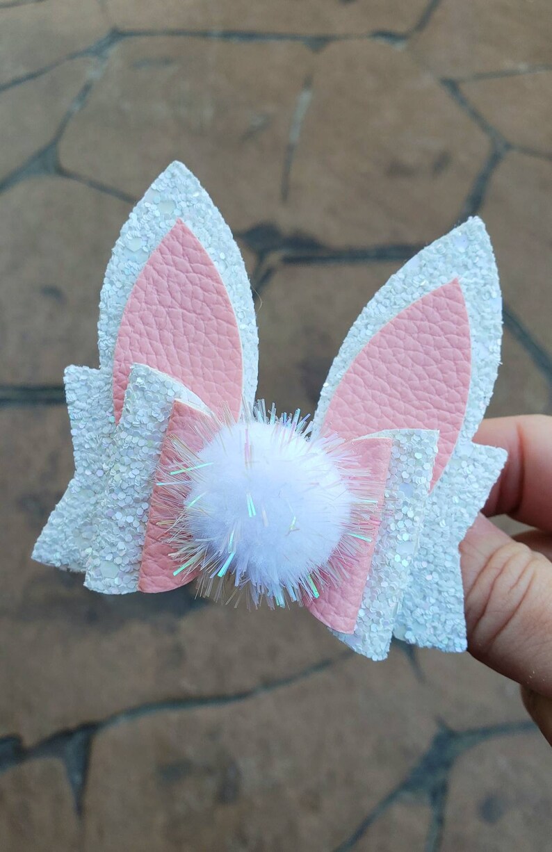 May include: A pink and white glitter bunny ear hair bow with a white pom-pom in the center.