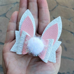 May include: A white and pink glitter hair bow with a white pom-pom in the center. The bow is shaped like bunny ears.