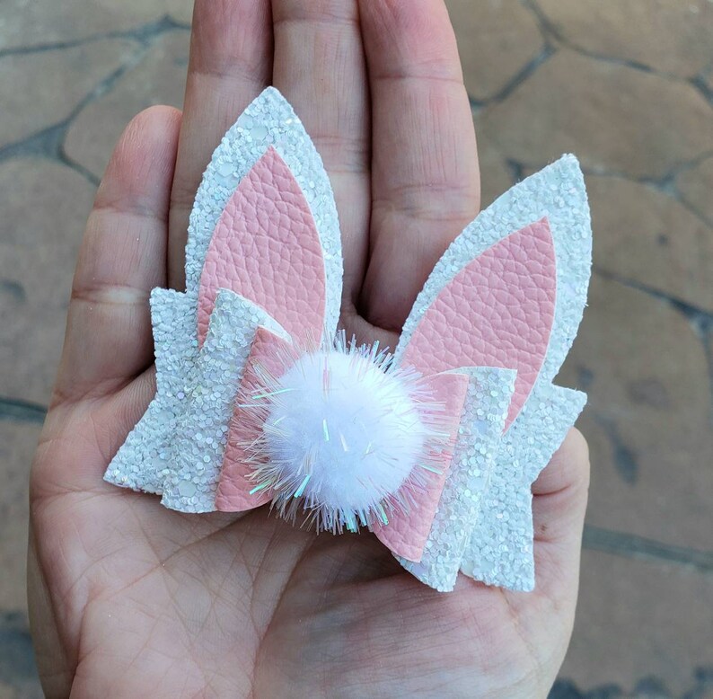May include: A white and pink glitter hair bow with a white pom-pom in the center. The bow is shaped like bunny ears.