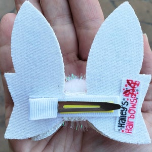 May include: White bunny ear hair clip with a white pom pom in the center and a yellow alligator clip. The clip has a white ribbon with the text "Hailey's Hairbows" in red and black.