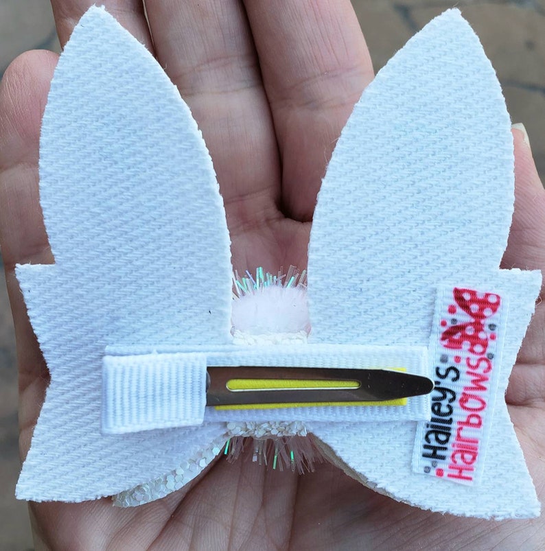 May include: White bunny ear hair clip with a white pom pom in the center and a yellow alligator clip. The clip has a white ribbon with the text "Hailey's Hairbows" in red and black.