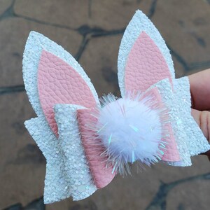 May include: A white and pink glitter bow with a white fluffy pom-pom in the center. The bow is made of faux leather and has a glittery finish.