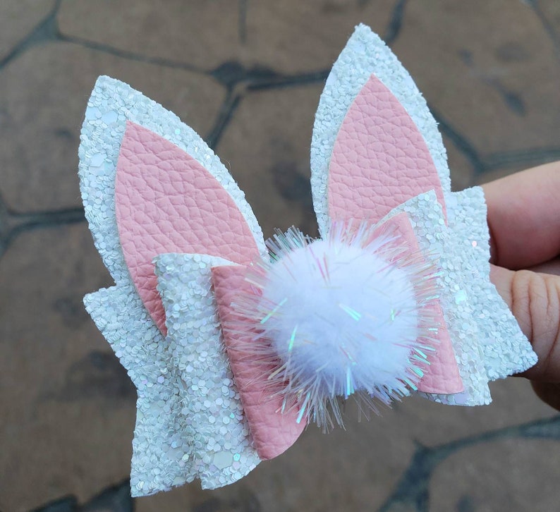 May include: A white and pink glitter bow with a white fluffy pom-pom in the center. The bow is made of faux leather and has a glittery finish.