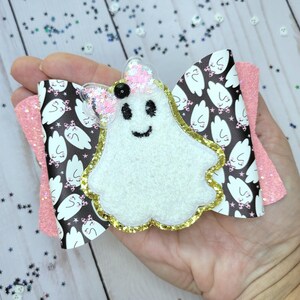 May include: A Halloween-themed hair bow featuring a white ghost with a smiling face, outlined in gold glitter. The bow has a black and white patterned center with pink glitter accents, perfect for festive hairstyles.