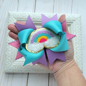 May include: A turquoise, purple, and pink hair bow with a rainbow and cloud applique. The applique is embroidered with a rainbow and a fluffy white cloud.