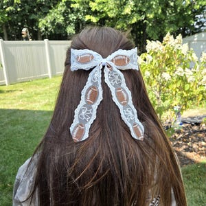 May include: A white lace hair bow featuring a repeating pattern of brown footballs and white outlined bows. The accessory has two long, dangling ribbon tails and a central knot, constructed from a delicate, semi-sheer white lace fabric.