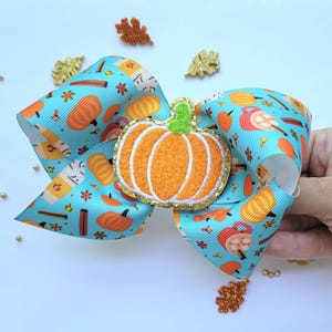 May include: A blue and white bow with a pumpkin design. The bow has a brown cinnamon stick, a red mug, and a pumpkin design. The bow is decorated with a felt pumpkin with a green stem.