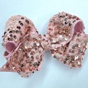 May include: A large, pink hair bow covered in rose gold sequins. The bow is made of a soft, velvety material and has a classic bow shape. The sequins are densely packed, giving the bow a sparkly appearance. The bow is likely for a child or for a special occasion.