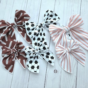 May include: Several fabric hair bows in various patterns. Two bows feature a brown football design with white stitching. Two bows have a black and white soccer ball pattern. Two bows have a white background with red stitching.