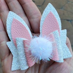 May include: A white and pink glitter bunny ear hair bow with a white pom-pom in the center.