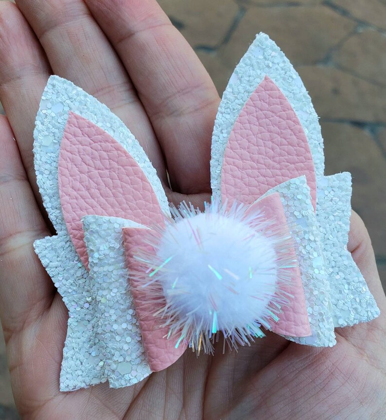 May include: A white and pink glitter bunny ear hair bow with a white pom-pom in the center.