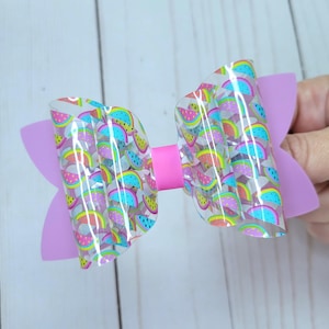 May include: A handmade hair bow featuring a watermelon pattern on a clear overlay. The bow has a light pink center and solid light pink wings. The watermelon slices are in various colors, including red, yellow, and green.
