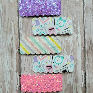 May include: Four hair clips with a back to school theme. Two clips are purple and pink glitter, one is striped in pastel colors, and one has a white background with a pattern of school supplies in blue, pink, and yellow.