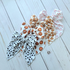 May include: Three white fabric hair bows with different sports-themed prints. The bows feature basketballs, soccer balls, and footballs.