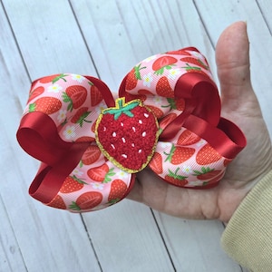 May include: A large, red and pink hair bow with a strawberry theme. The bow features a repeating pattern of strawberries on a pink background, with solid red ribbon accents. A detailed, embroidered strawberry is centered on the bow.