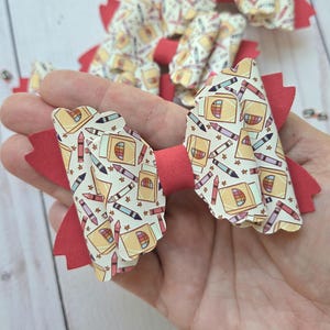 May include: A hair bow featuring a repeating pattern of yellow crayon boxes and colorful crayons on a white background, accented with a solid red base layer and a red center band. The edges of the bow have a scalloped, decorative finish.
