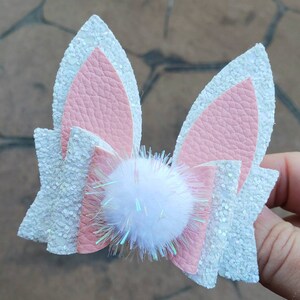 May include: White and pink glitter bunny ears with a white pom-pom in the center.