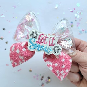 May include: A pink and white glitter bow with a clear plastic center and a white embroidered patch that says "Let it Snow".